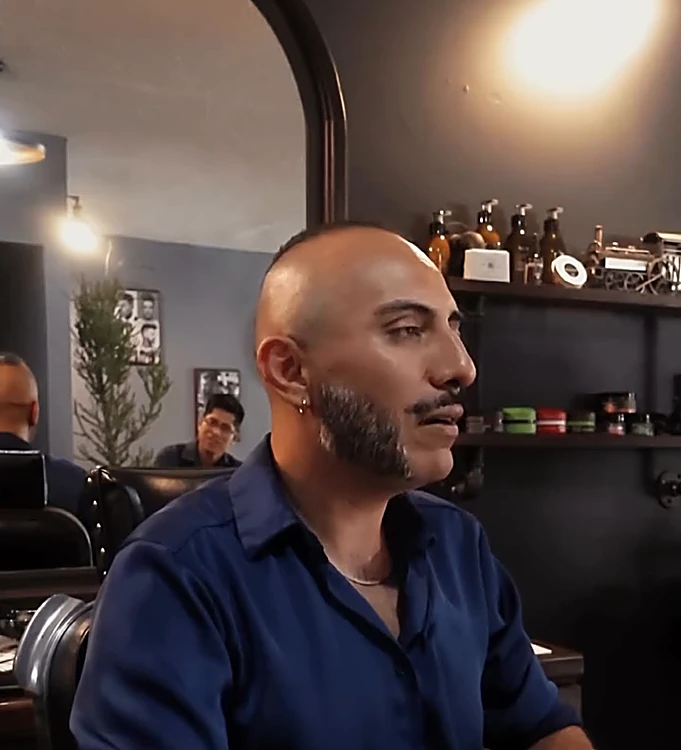 Carlos Fernández, de cabeza rapada, barba y camisa azul marino, sentado en la barbería