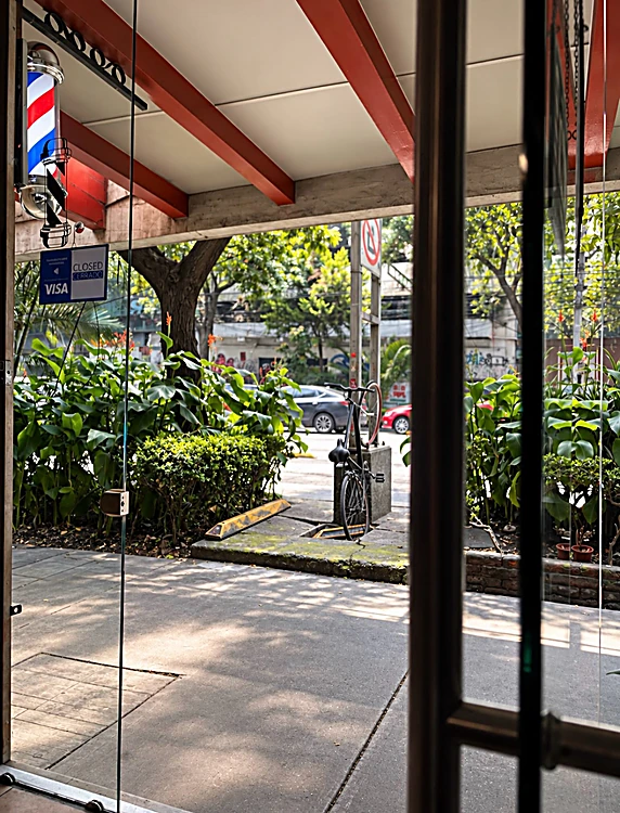 Vista desde la puerta hacia Av. Yucatán: vigas rojas de la marquesina, poste de barbero y árboles de la calle