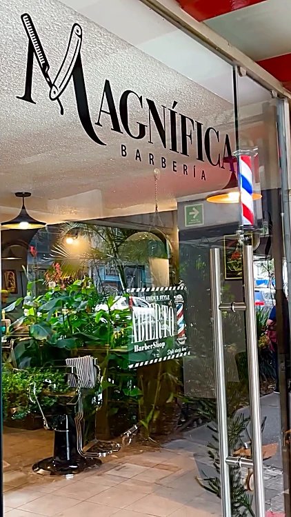 Vidrio de la entrada con el logo de Magnífica Barbería, el poste de barbero y plantas, con el salón al fondo