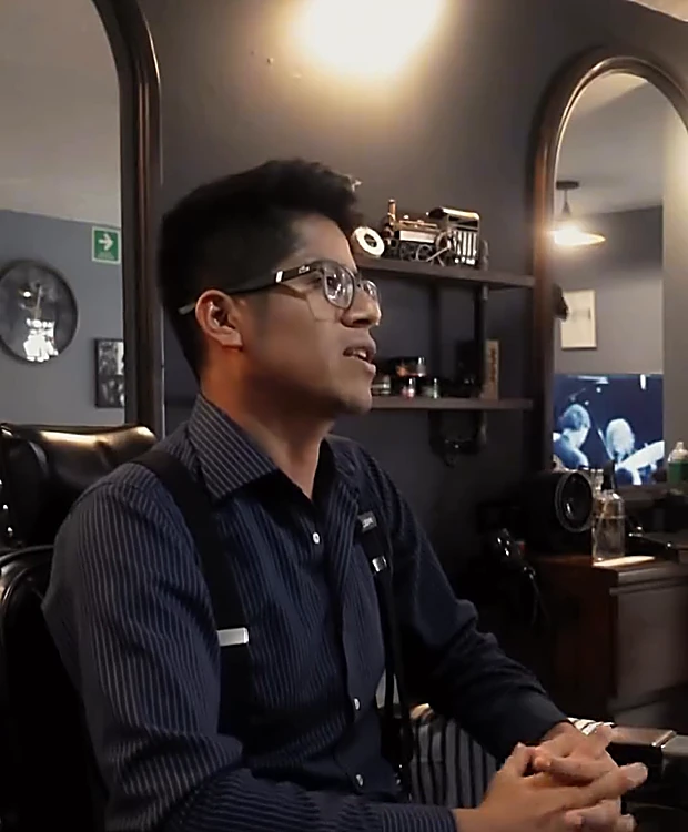 Rubén Mancilla, de lentes y camisa oscura, sentado en un sillón de la barbería frente a los espejos de arco